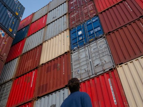 man in black jacket standing in front of red and blue intermodal containers